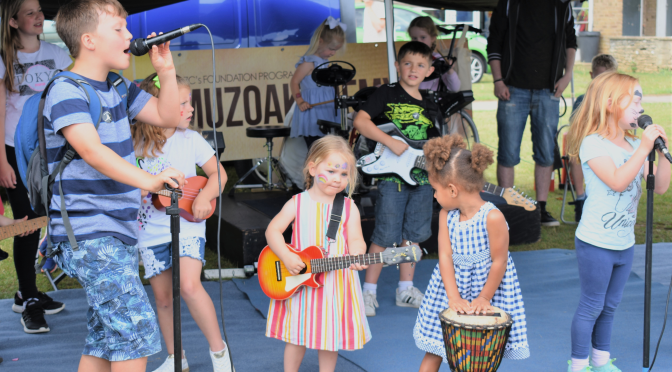 A group of kids of varying ages stand under a tent and play instruments together. One older child is passionately singing into a microphone.